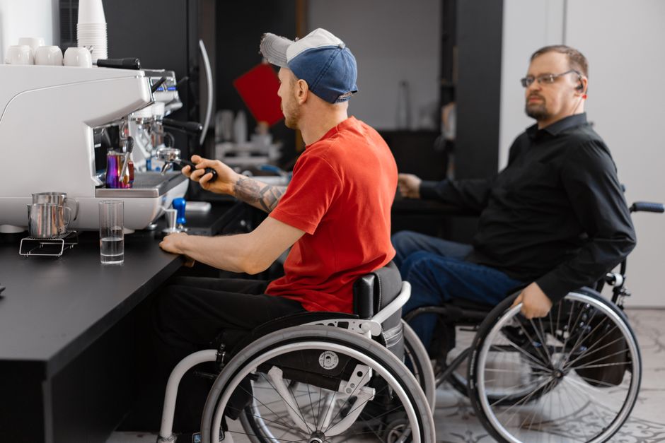 Two men in wheelchairs work at a coffee shop, showcasing inclusivity in the workplace.